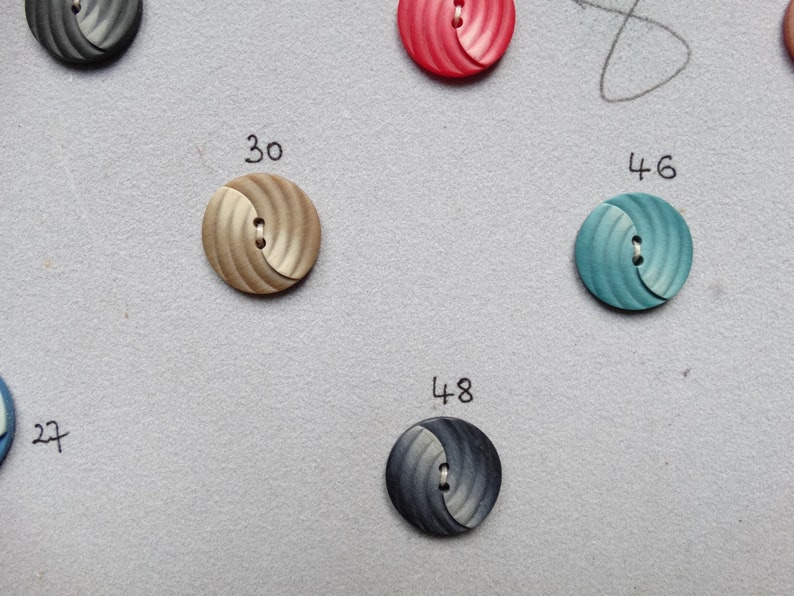 May include: Close-up of several round buttons in various colors, including tan, teal, and navy. Each button has a swirled design and two holes for sewing. Numbers 27, 30, 46, and 48 are written near the buttons.