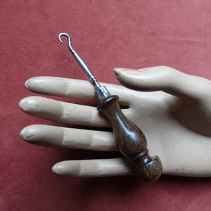 May include: A vintage crochet hook with a polished wooden handle and a silver metal hook. The hook is held in a mannequin hand, showcasing its design and craftsmanship. The hook is a tool for crafting.
