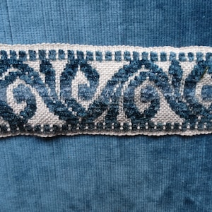 May include: A close-up of a blue velvet fabric with a white and blue trim. The trim features a repeating pattern of stylized swirls.