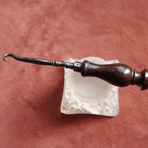 May include: A dark wooden handle with a metal hook tool, resting on a frosted glass container. The hook is curved and the handle is a rich, dark brown color. The tool appears to be vintage or antique.