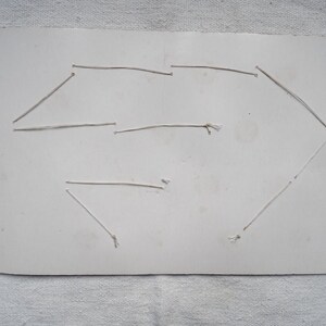 May include: A rectangular, off-white card with a geometric design created with thin, light-colored string. The string is attached to the card with small knots, forming a shape. The card has some discoloration.
