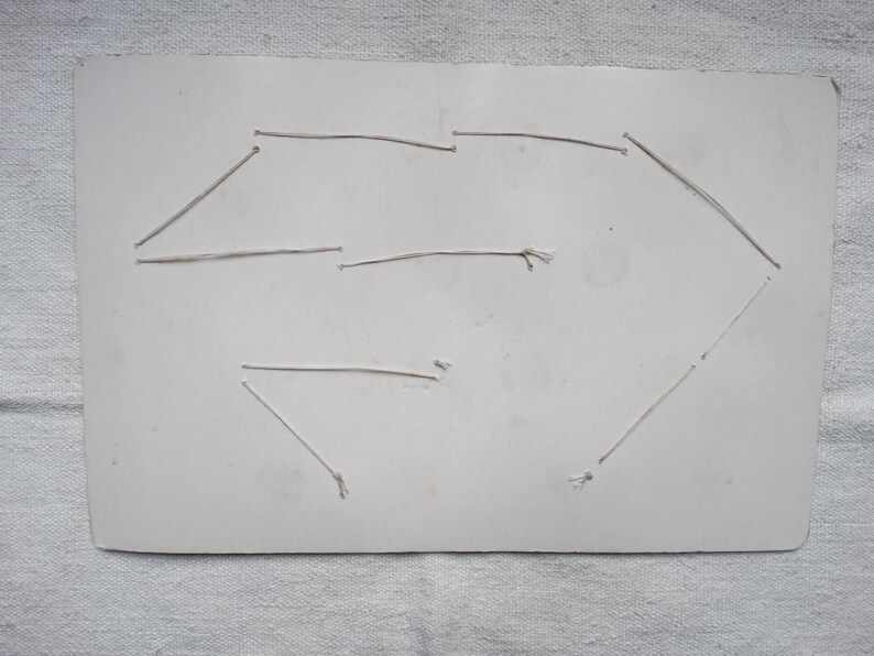 May include: A rectangular, off-white card with a geometric design created with thin, light-colored string. The string is attached to the card with small knots, forming a shape. The card has some discoloration.
