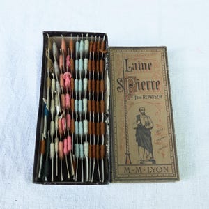 Op de afbeelding: Een open, vintage naaidoos met een verscheidenheid aan kleurrijke garenspoelen. De zwarte doos heeft een bruin label met de tekst "Laine Pierre" en een illustratie van een man. De spoelen zijn in tinten roze, blauw, bruin en crème.