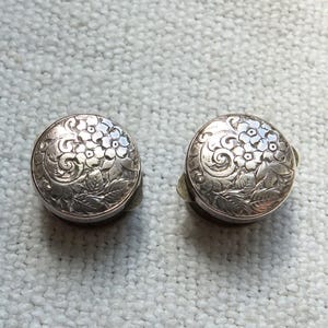 Könnte beinhalten: Zwei silberfarbene Manschettenknöpfe mit einem filigranen Blumendesign. Die Manschettenknöpfe sind rund und haben ein erhabenes Design aus Blumen und Blättern.