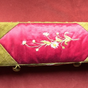 May include: Rectangular jewelry box with a red satin center panel embroidered with white flowers and green leaves. The box has olive green suede sides and gold trim. Small gold feet are visible.