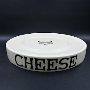 May include: A round, off-white ceramic cheese plate with a raised edge. The word CHEESE is printed in large, bold black capital letters along the side. The center features a small black graphic logo with the text JOANNE HUDSON PHILADELPHIA.