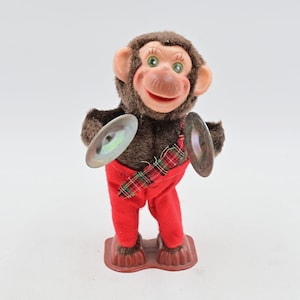 May include: A vintage-style mechanical monkey toy covered in brown synthetic fur with a plastic face, green eyes, and a wide smile. The monkey wears bright red trousers and a red, green, and black plaid vest, holding two metallic cymbals.