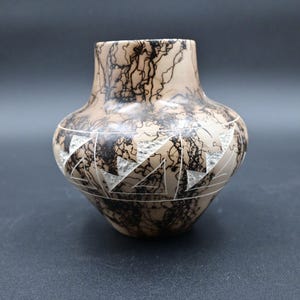 May include: A beige ceramic vase with a rounded body and a slightly flared rim. The vase features a black, lightning-like pattern across the surface. A band of white, geometric shapes encircles the middle of the vase, adding a decorative element.