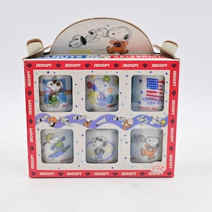 May include: A Peanuts Celebration boxed set containing six white ceramic mugs. Each mug features a unique Snoopy graphic, including space-themed designs, an American flag, and various outfits. The box is red and white with Snoopy branding.