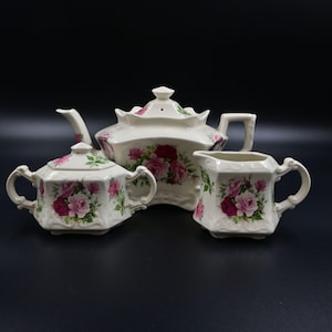 May include: A cream-colored ceramic tea set consisting of a teapot with a lid, a lidded sugar bowl with two handles, and a creamer pitcher. Each piece features a scalloped edge design and a floral pattern of pink and deep red roses with green leaves.