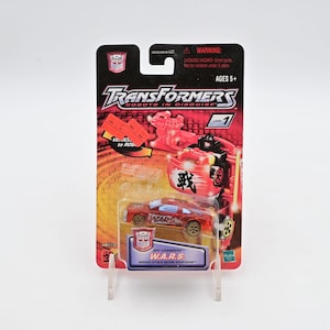 May include: A red and gold Transformers toy car in its original packaging. The packaging features the Transformers logo, the text "ROBOTS IN DISGUISE", and the name "W.A.R.S." The toy is a "Wicked Attack Recon Sportscar" and is for ages 5+.