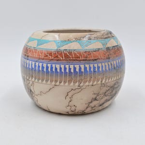 May include: A round, cream-colored ceramic vase with a decorative pattern. The vase features bands of turquoise, red, and blue geometric designs. The lower portion has a unique crackle pattern. A handcrafted piece.