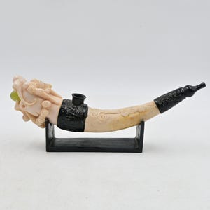May include: A curved smoking pipe featuring a carved off-white dragon head at the bowl end, holding a small green sphere in its mouth. The pipe has black decorative metal bands at the bowl and mouthpiece, resting on a black rectangular stand.