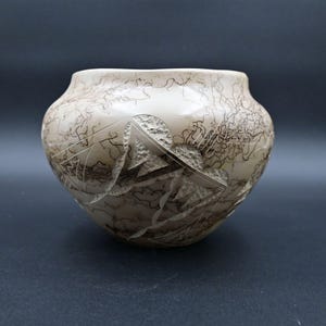 May include: A cream-colored ceramic bowl with a wide opening and rounded body. The bowl features etched mountain and abstract designs, with dark brown lines and textured areas. The bowl is approximately 6 inches in diameter.