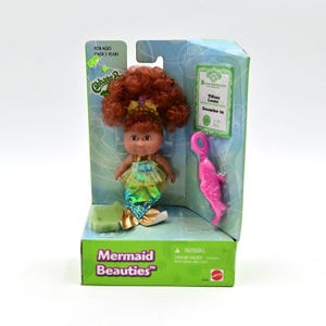 May include: A Mermaid Beauties doll in its original packaging. The doll has red curly hair, a mermaid tail, and a pink accessory. The packaging includes the text "FOR AGES OVER 3 YEARS" and "Mermaid Beauties".
