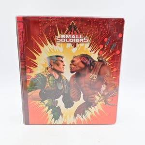 May include: A rectangular Small Soldiers movie case featuring a red background with a circuit board pattern. The front displays a soldier in camouflage and a brown creature facing each other against a yellow burst, with the movie title at the top.
