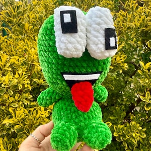 Peut inclure: Peluche grenouille verte faite à la main, avec de grands yeux blancs et des pupilles noires. La grenouille a une langue rouge qui dépasse. La peluche est tenue devant un fond de feuillage jaune et vert. Jouet doux et fantaisiste.