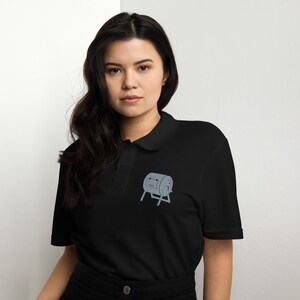 May include: Black polo shirt featuring a grey cartoon drum graphic embroidered on the chest. The shirt has a collared neckline and short sleeves. The drum graphic has a smiling face.