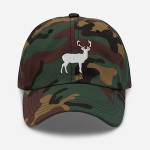 Peut inclure: Casquette de baseball camouflage avec une silhouette de cerf blanche brodée sur le devant. La casquette a une visière incurvée et une sangle réglable à l'arrière. Le motif de la casquette comprend des nuances de vert, de marron et de beige.
