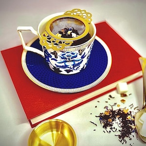 May include: A white teacup with a blue and gold floral design, a gold tea strainer, and a blue and white saucer. The teacup and saucer are sitting on a red book. Loose leaf tea is scattered around the teacup and saucer.