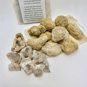 May include: A collection of tan, rough-textured geode rocks, some intact and others broken to reveal white crystalline interiors. A beige drawstring fabric bag is included, featuring printed instructions titled How To Break A Geode.