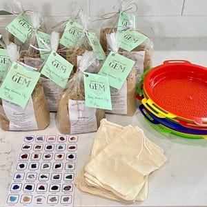 May include: A collection of clear plastic bags filled with sand and gemstones, each with a light green tag labeled "you're a real GEM" and "Happy Valentine's Day". Includes a stack of colorful plastic sifters, a gemstone identification chart, and several small beige drawstring pouches.