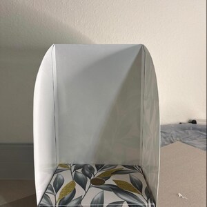 May include: A white, U-shaped display stand featuring a base with a botanical pattern of grey and white leaves accented with gold glitter. The structure has a flat, rectangular bottom and two curved side panels that extend upward to create a backdrop.
