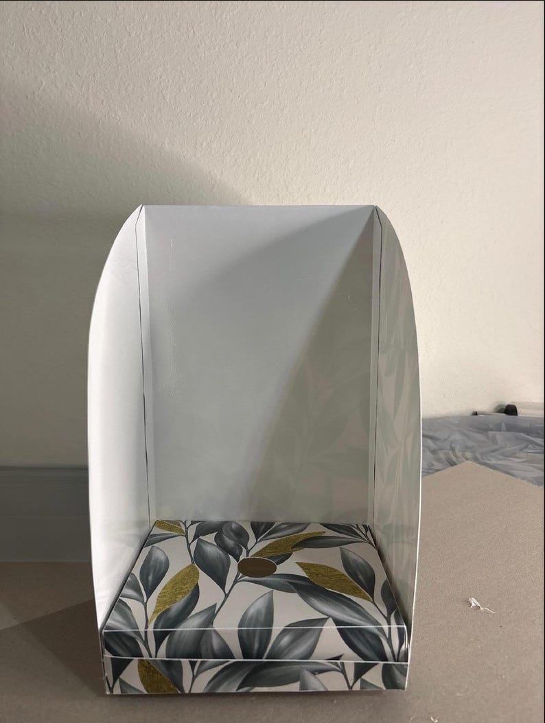 May include: A white, U-shaped display stand featuring a base with a botanical pattern of grey and white leaves accented with gold glitter. The structure has a flat, rectangular bottom and two curved side panels that extend upward to create a backdrop.