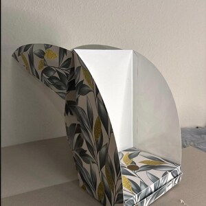 May include: A semi-open cardboard display box featuring a botanical pattern of dark gray leaves and gold glitter accents on a white background. The structure has a curved side panel and a flat base, with a plain white interior lining visible.