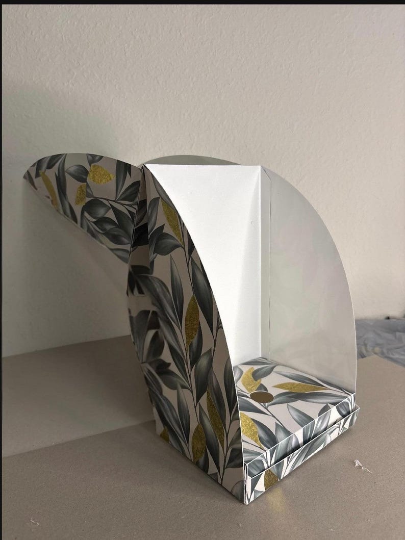 May include: A semi-open cardboard display box featuring a botanical pattern of dark gray leaves and gold glitter accents on a white background. The structure has a curved side panel and a flat base, with a plain white interior lining visible.