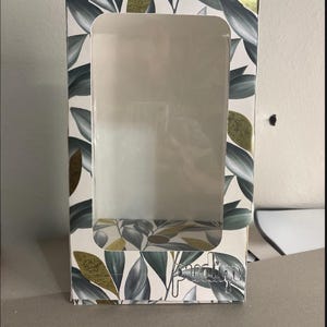 May include: A rectangular cardboard display box featuring a large clear plastic window on the front. The exterior is decorated with a pattern of dark green leaves and gold foil accents. The word pudim is printed in a silver metallic font at the bottom.