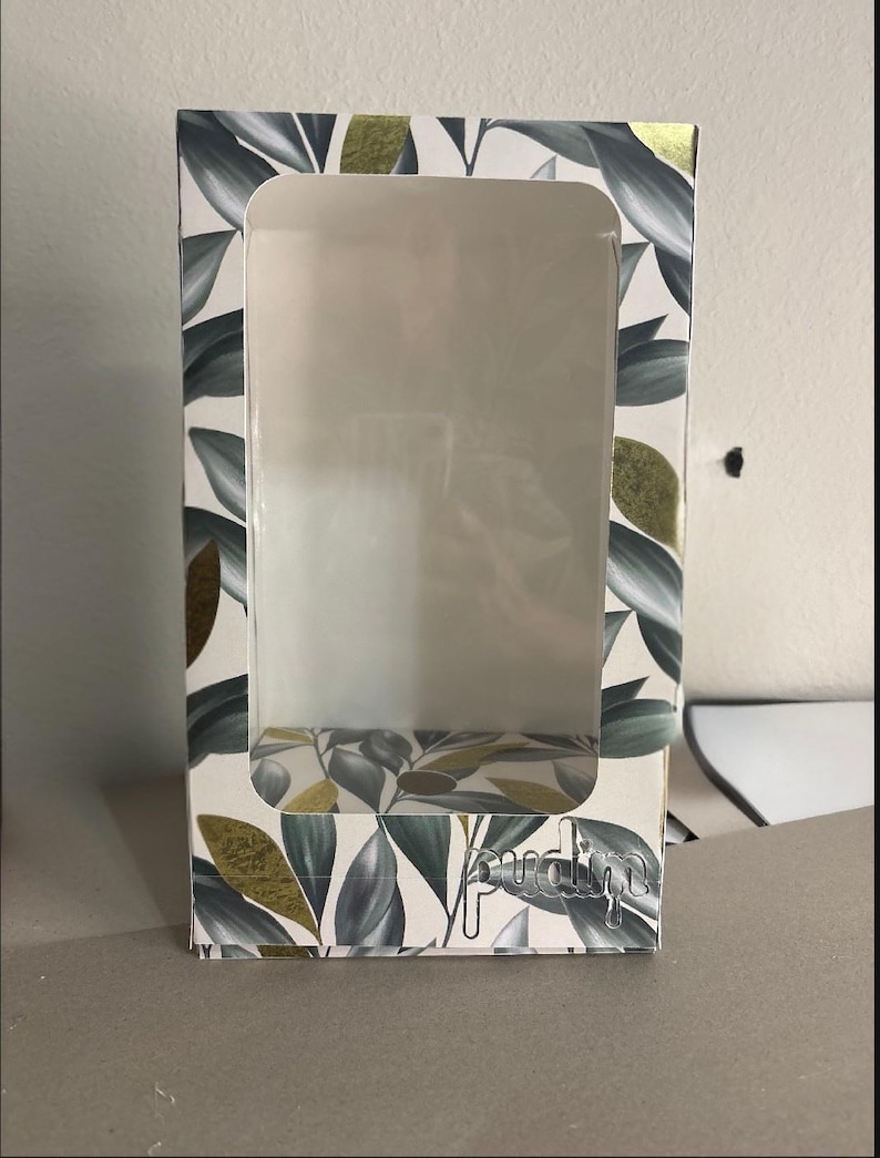 May include: A rectangular cardboard display box featuring a large clear plastic window on the front. The exterior is decorated with a pattern of dark green leaves and gold foil accents. The word pudim is printed in a silver metallic font at the bottom.