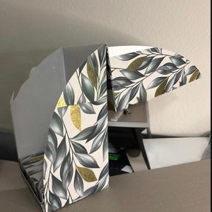 May include: A partially assembled cardboard box featuring a botanical pattern of dark green and grey leaves on a cream background, accented with scattered gold foil leaf shapes. The interior of the box is plain white with visible folding flaps.
