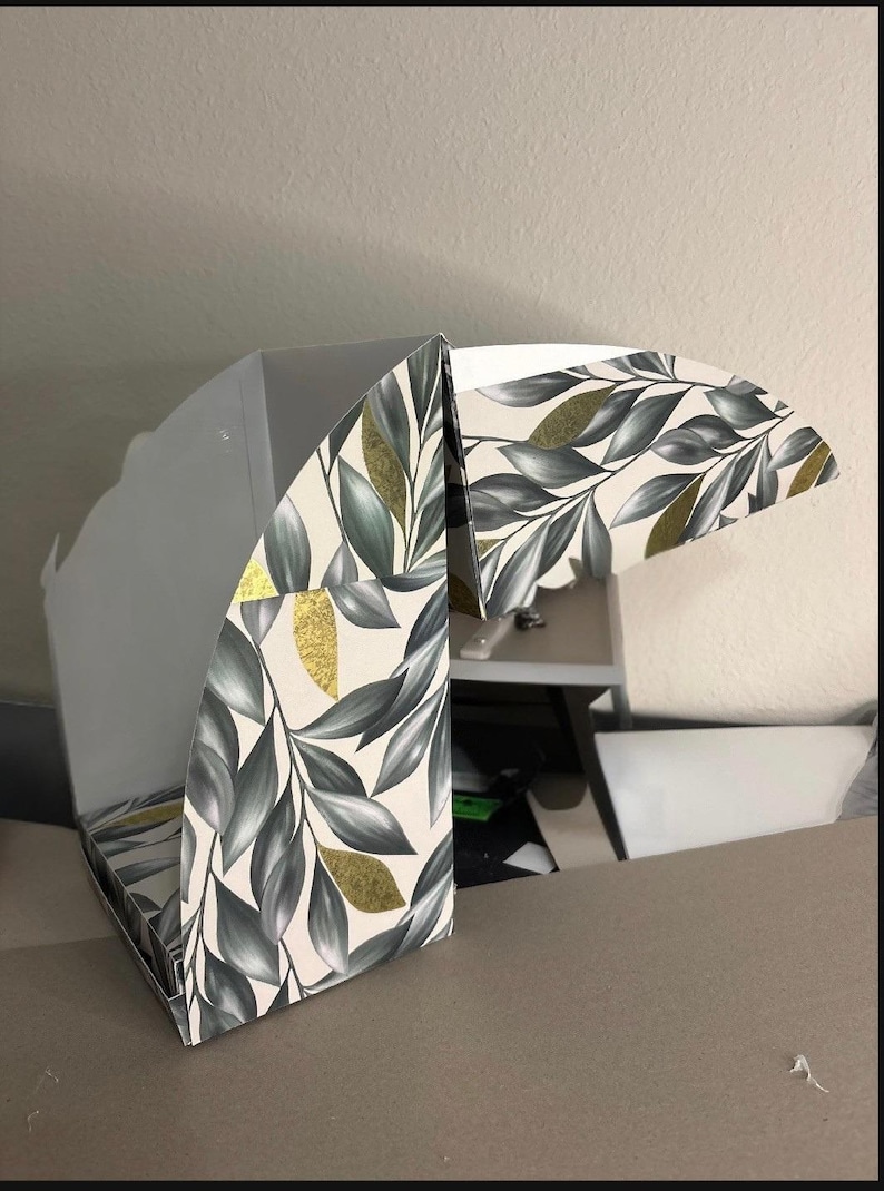 May include: A partially assembled cardboard box featuring a botanical pattern of dark green and grey leaves on a cream background, accented with scattered gold foil leaf shapes. The interior of the box is plain white with visible folding flaps.