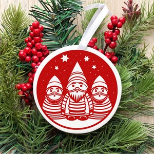 May include: A red and white Christmas ornament featuring three white gnomes with red hats and beards. The gnomes are standing in a row with stars in the background.