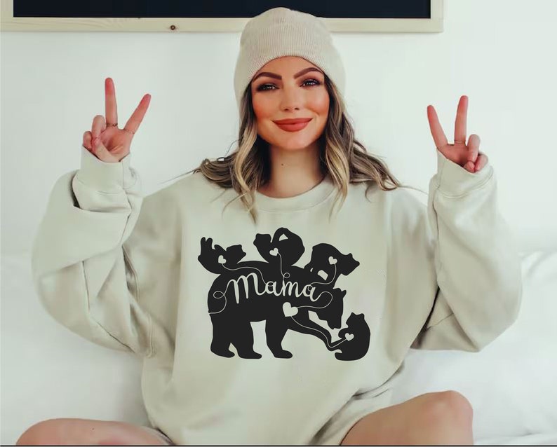 Puede incluir: Sudadera beige con un gr&aacute;fico negro de un oso y cachorros, con la palabra "Mama" en cursiva. El dise&ntilde;o del oso incluye detalles de coraz&oacute;n. La persona de la imagen lleva un gorro beige.