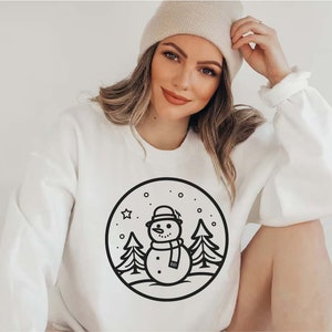 May include: White sweatshirt with a black and white illustration of a snowman in a winter scene. The snowman is wearing a hat and scarf. There are trees and snowflakes in the background.
