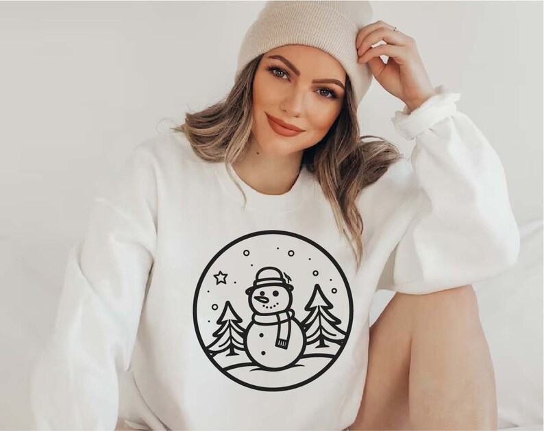 May include: White sweatshirt with a black and white illustration of a snowman in a winter scene. The snowman is wearing a hat and scarf. There are trees and snowflakes in the background.