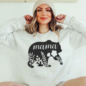 Puede incluir: Sudadera gris claro con un dise&ntilde;o de oso negro. La silueta del oso tiene la palabra "mama" en escritura blanca y est&aacute; decorada con patrones florales y de hojas. La sudadera es de estilo cuello redondo.