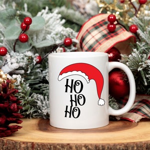 May include: White ceramic mug featuring a red Santa hat design and the text "HO HO HO" in black. The mug is surrounded by Christmas decorations, including pine branches, red berries, and a plaid ribbon.