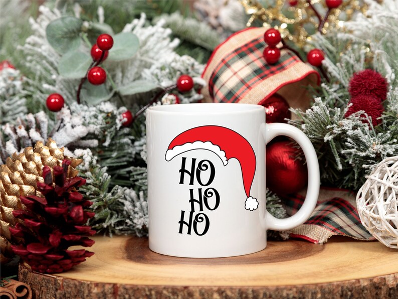 May include: White ceramic mug featuring a red Santa hat design and the text "HO HO HO" in black. The mug is surrounded by Christmas decorations, including pine branches, red berries, and a plaid ribbon.