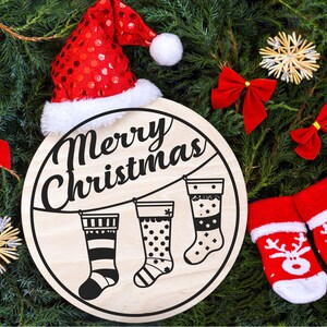May include: A round wooden sign with the text "Merry Christmas" in black script. Three black and white stockings are hanging from a string with clothespins. The sign is surrounded by green pine branches, red bows, and a red Santa hat.