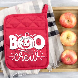 Peut inclure: Une manique de four rouge matelass&eacute;e avec du texte blanc qui dit "Boo Crew" et une citrouille avec un visage de citrouille. Il y a un fouet et une petite chauve-souris au-dessus de la citrouille. La manique est sur un tissu &agrave; carreaux avec trois pommes rouges dans un plateau en bois.