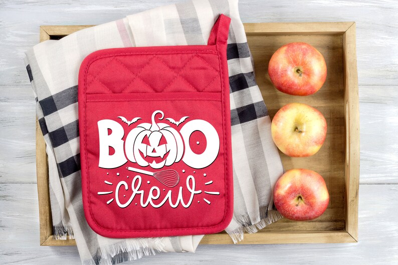 Peut inclure: Une manique de four rouge matelass&eacute;e avec du texte blanc qui dit "Boo Crew" et une citrouille avec un visage de citrouille. Il y a un fouet et une petite chauve-souris au-dessus de la citrouille. La manique est sur un tissu &agrave; carreaux avec trois pommes rouges dans un plateau en bois.