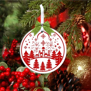 May include: White Christmas ornament with a red silhouette of a town with trees and fireworks. The ornament is hanging from a green ribbon with a bow.