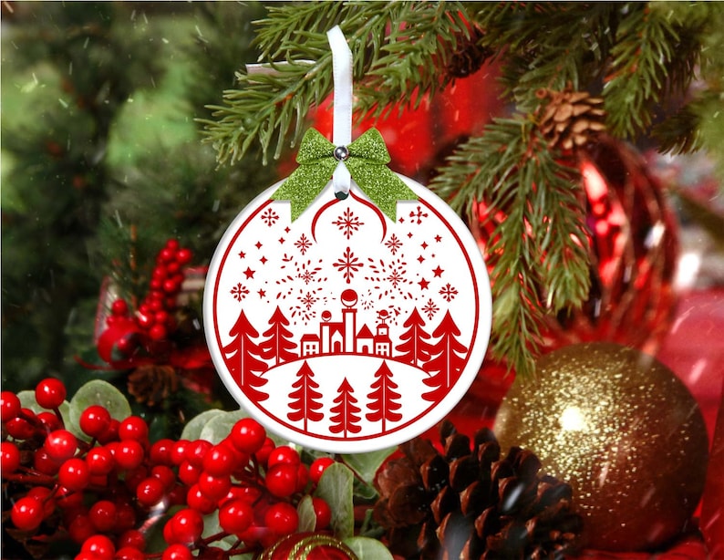 May include: White Christmas ornament with a red silhouette of a town with trees and fireworks. The ornament is hanging from a green ribbon with a bow.