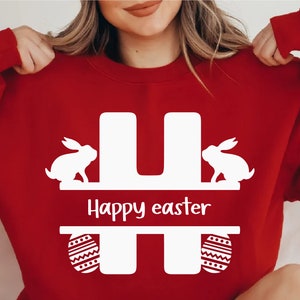 Puede incluir: Sudadera roja con una letra H blanca, dos conejos blancos y el texto "Happy easter" en la parte delantera.