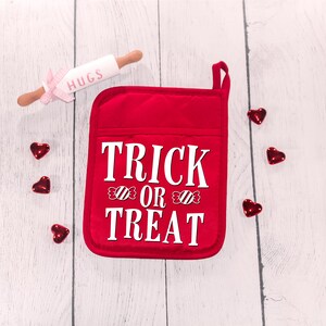 Peut inclure: Une manique de four rouge avec le texte "TRICK OR TREAT" en blanc avec des motifs de cannes en sucre. La manique est sur un fond de bois blanc avec des c&oelig;urs rouges &eacute;parpill&eacute;s autour.