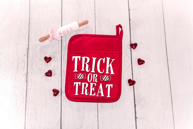 Peut inclure: Une manique de four rouge avec le texte "TRICK OR TREAT" en blanc avec des motifs de cannes en sucre. La manique est sur un fond de bois blanc avec des c&oelig;urs rouges &eacute;parpill&eacute;s autour.