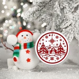 May include: A white ceramic Christmas ornament with a red design of a house, two pine trees, and a star in a snowy landscape.